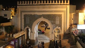 A historic gate with intricate designs, leading to a street with parked cars and pedestrians. - Dar Fatimazahra (Fes)