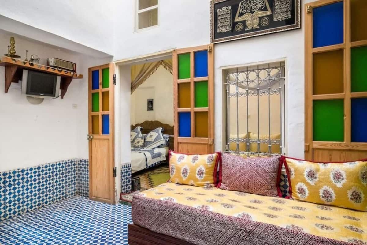 Une chambre avec un lit, un téléviseur fixé au mur et une fenêtre ornée de grilles décoratives.
