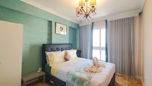 A neatly made bed with a duvet, a bedside table, a bench, a chandelier, and a window with curtains. -  Beautiful sea view Apt,in the heart of Alexandria (alexandria)