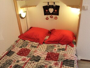 Une petite pièce accueillante avec un lit agrémenté d’une courtepointe matelassée et de deux oreillers rouges. On peut voir une tête de lit en bois avec un miroir et des lampes fixées au mur.