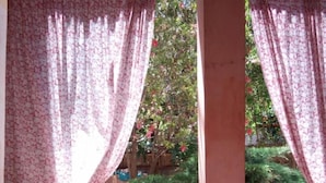 Una habitación con una mesa, una silla y un ventanal con cortinas florales, con vista a la vegetación exterior.