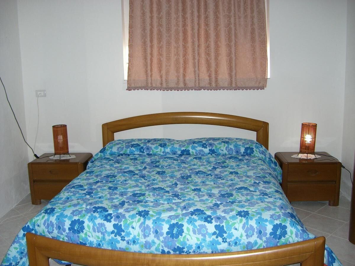 Une chambre à coucher avec un lit en bois, une couvre-lit à motifs floraux, deux tables de chevet avec des lampes et une fenêtre avec un rideau.