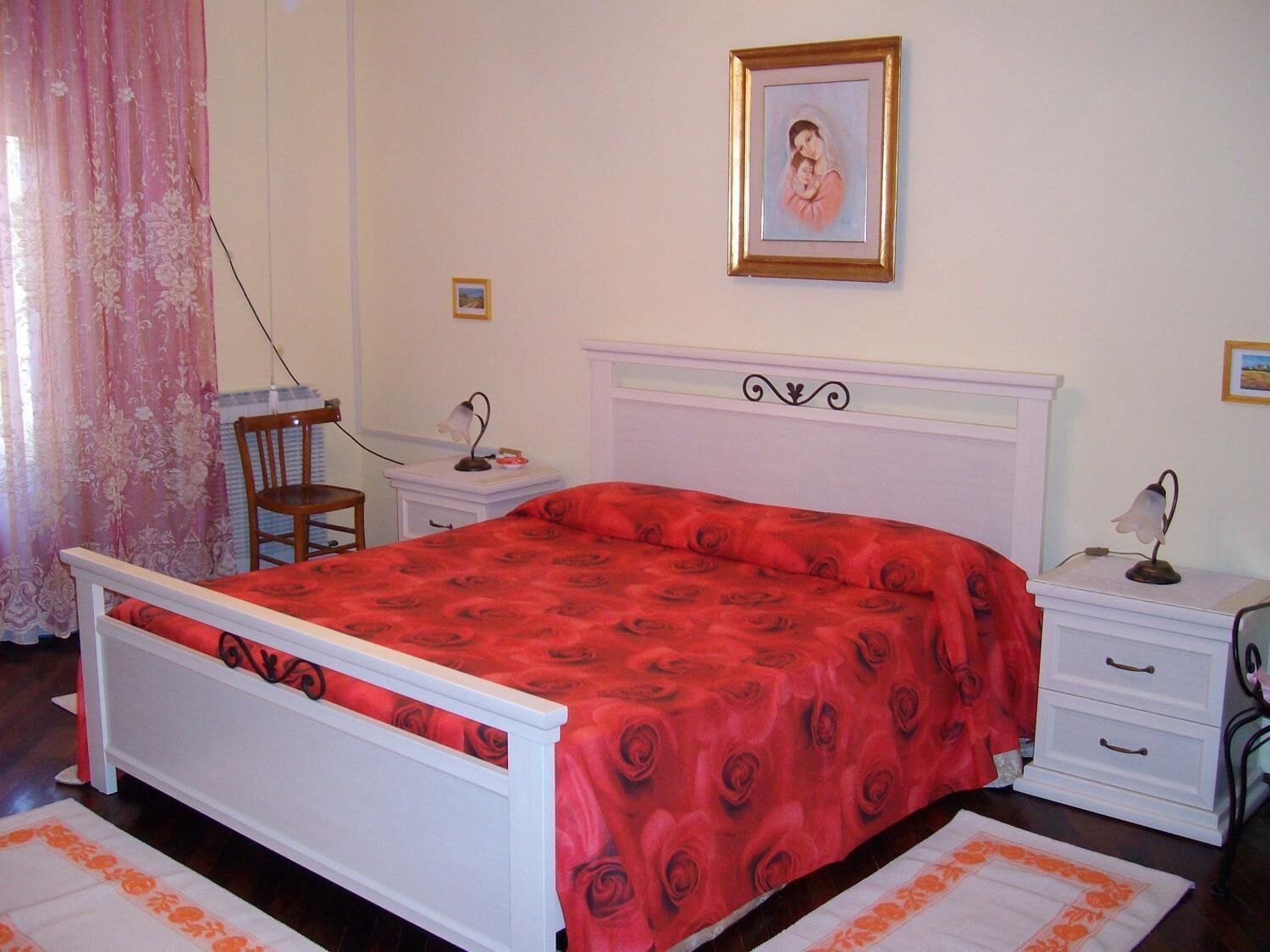 Una camera da letto con un letto bianco, una coperta con motivo floreale a rose rosse, un comodino con una lampada, una sedia e un quadro incorniciato appeso al muro.