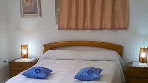 Un dormitorio con una cama, dos lámparas de noche, un ventanal con cortina, un cuadro en la pared y una alfombra amarilla.