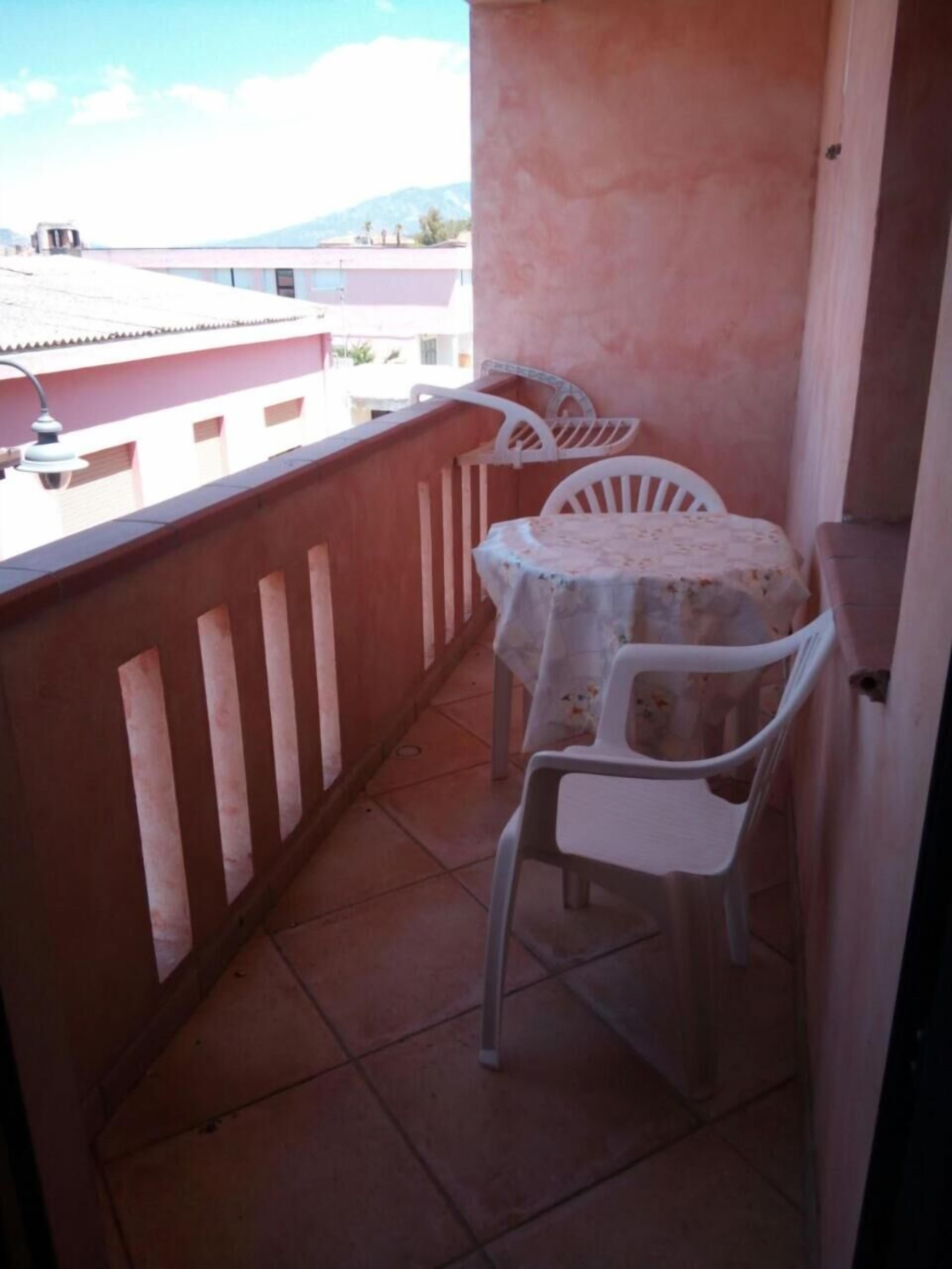 Un pequeño balcón con una mesa, dos sillas, barandilla y vistas a edificios y montañas al fondo.