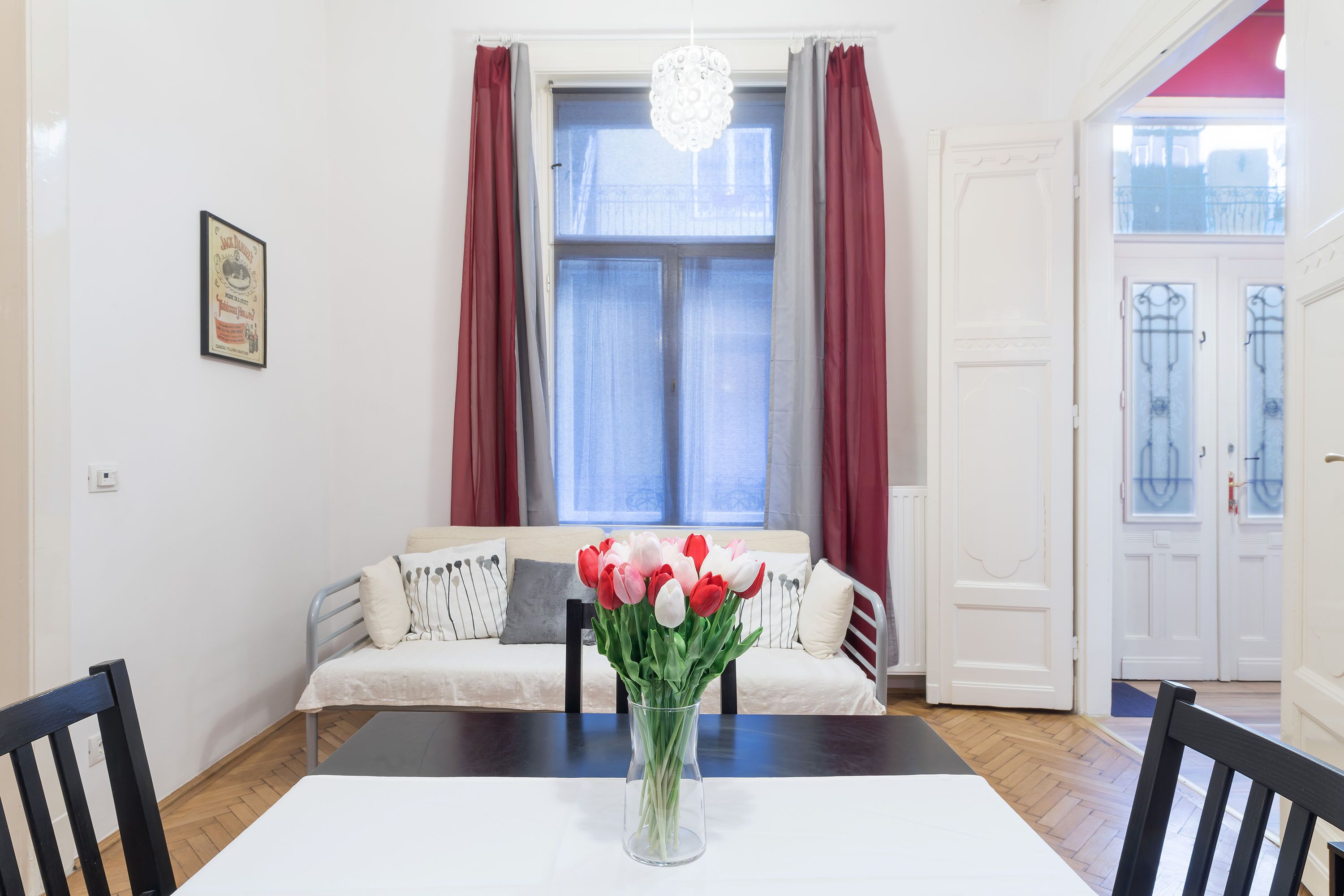 A dining area with a table, chairs, a vase of flowers, a sofa with cushions, a window with curtains, and a door.
