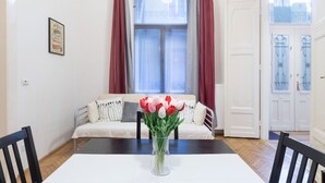 A dining area with a table, chairs, a vase of flowers, a sofa with cushions, a window with curtains, and a door. - Akacfa 18 Apartment (Budapest)