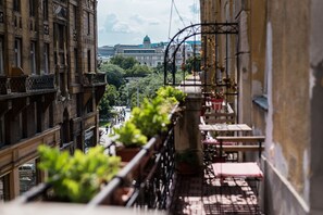 Un balcon agrémenté de plantes en pot, une table et des chaises, donnant sur une rue bordée d’immeubles et sur laquelle on aperçoit, au loin, une structure en forme de dôme.