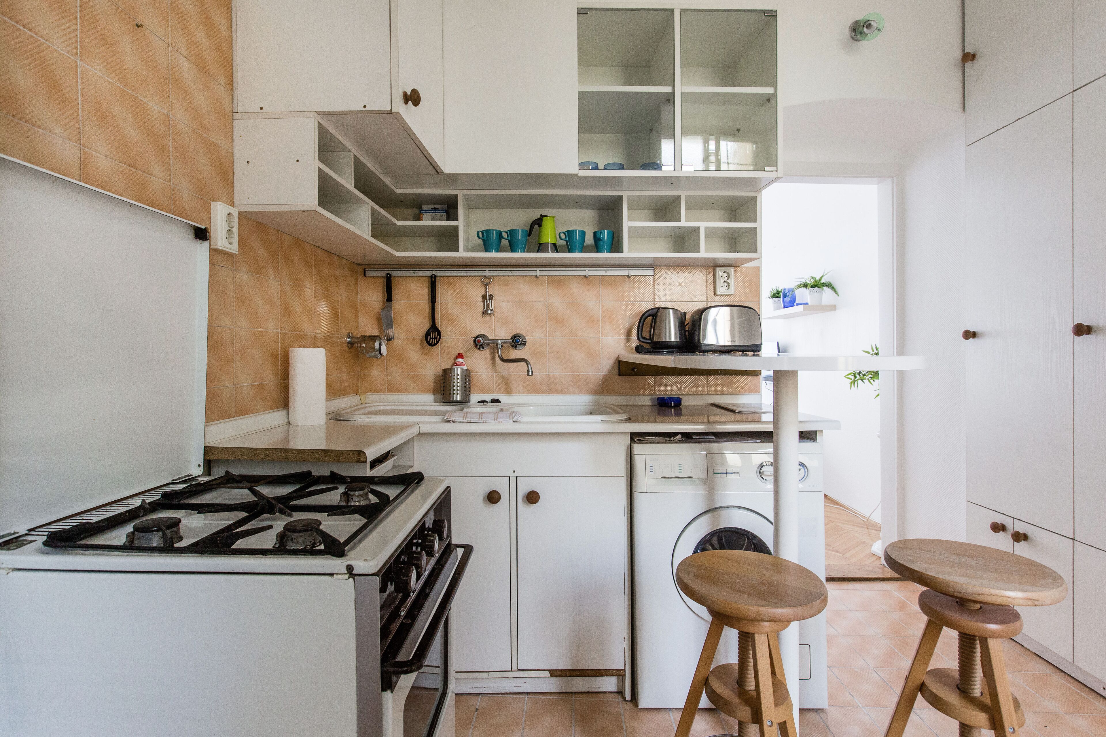 Een kleine keuken met een gasfornuis, een wasmachine en houten krukjes.
