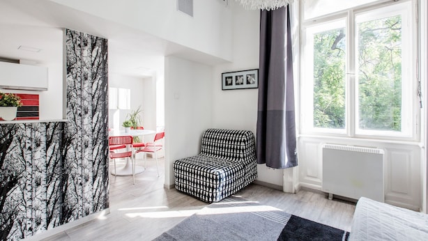 Uma sala de estar moderna com um sofá estampado, uma área de jantar com mesa vermelha e branca, e uma janela grande com vista para árvores.