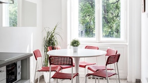 Um quarto claro com uma mesa redonda, cadeiras vermelhas, um vaso de planta e uma janela com vista para árvores.