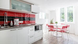 Cozinha moderna com armários brancos, painel de parede em vermelho e preto e uma cafeteira. Há uma área de jantar com mesa branca e cadeiras vermelhas.