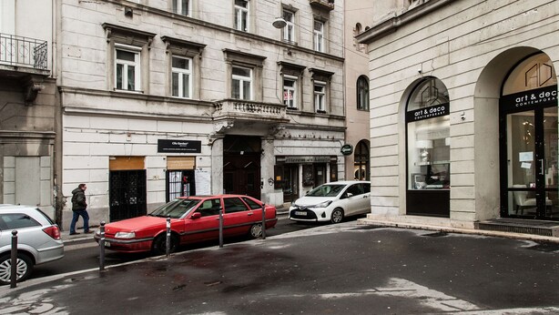 A street view with a building featuring multiple windows and balconies, a storefront with signage, and parked cars.