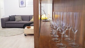 A room with a wooden cabinet containing multiple wine glasses, a grey sofa with cushions, a small wooden table, and a lamp in the background. - Harsfa 18 Apartment (Budapest)