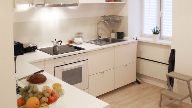 A modern kitchen with white cabinets, a built-in oven, a sink, and a window with blinds. - Harsfa 18 Apartment (Budapest)