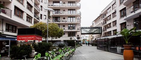 A row of green bicycles parked on a tiled sidewalk in front of a building with multiple balconies and a sign for a health club.