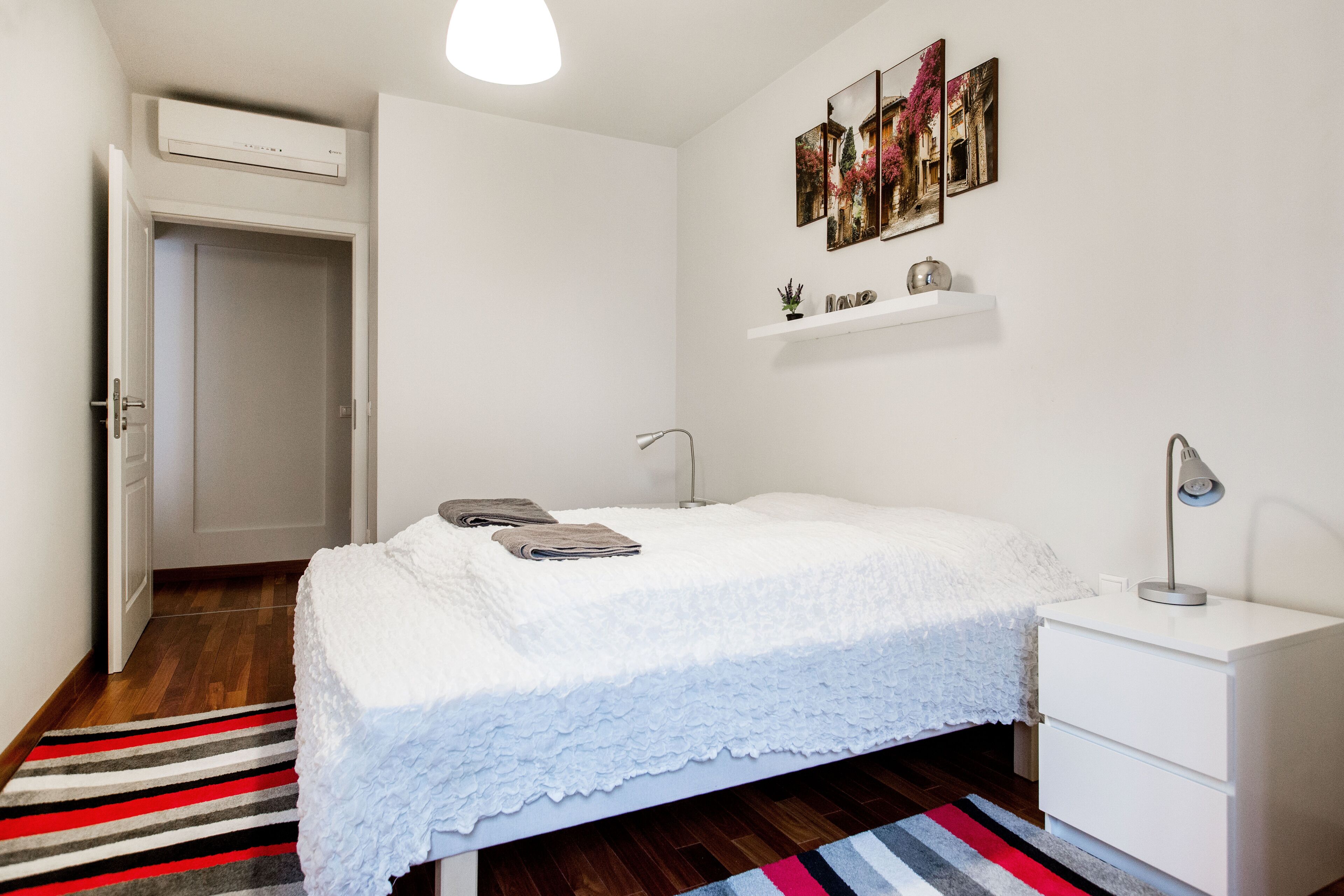 Un dormitorio con cama, mesitas de noche, un armario, un estante colgante con adornos y una puerta a otra habitación.