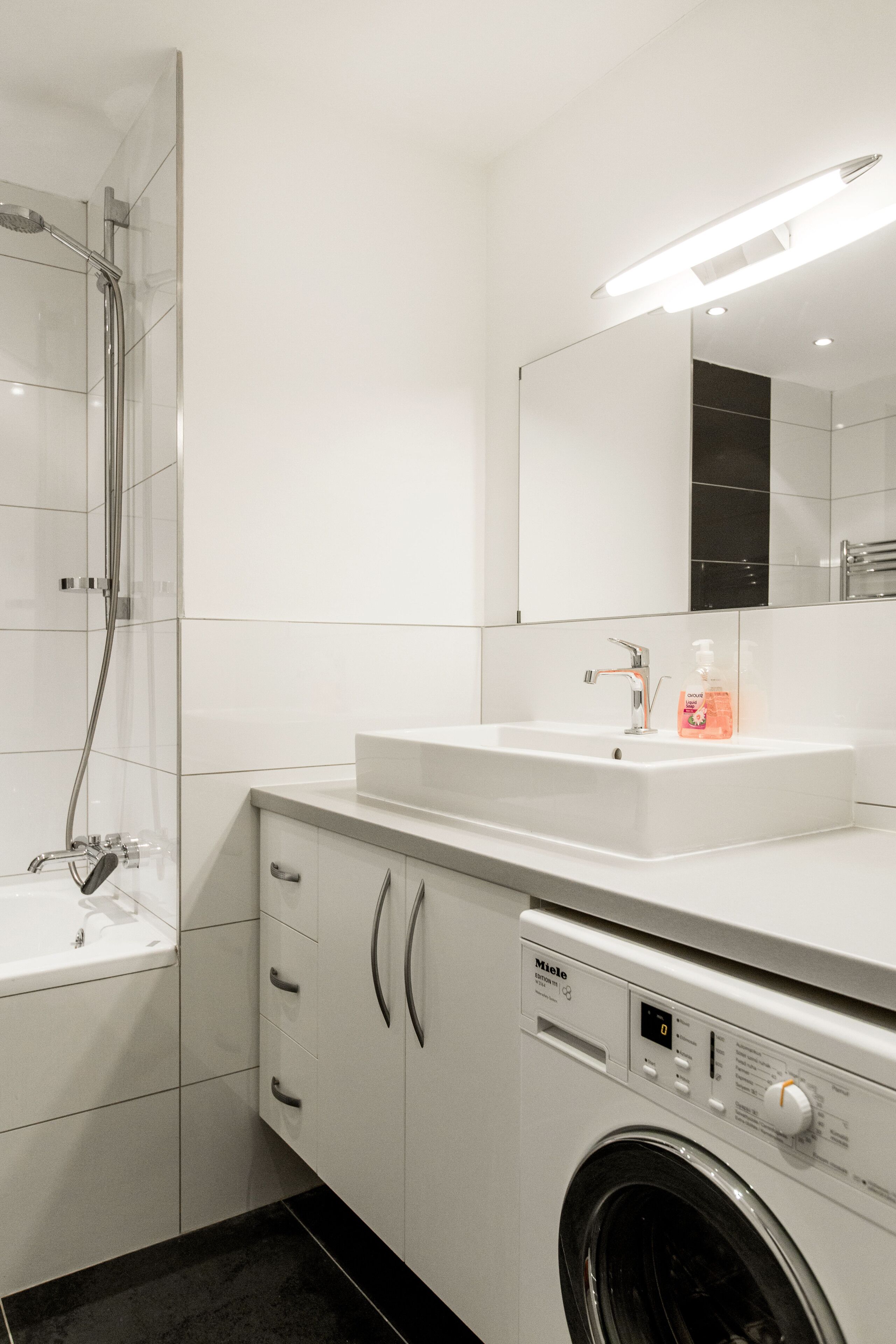 A modern bathroom with a glass-enclosed shower, a sink with a soap dispenser, and a front-loading washing machine.