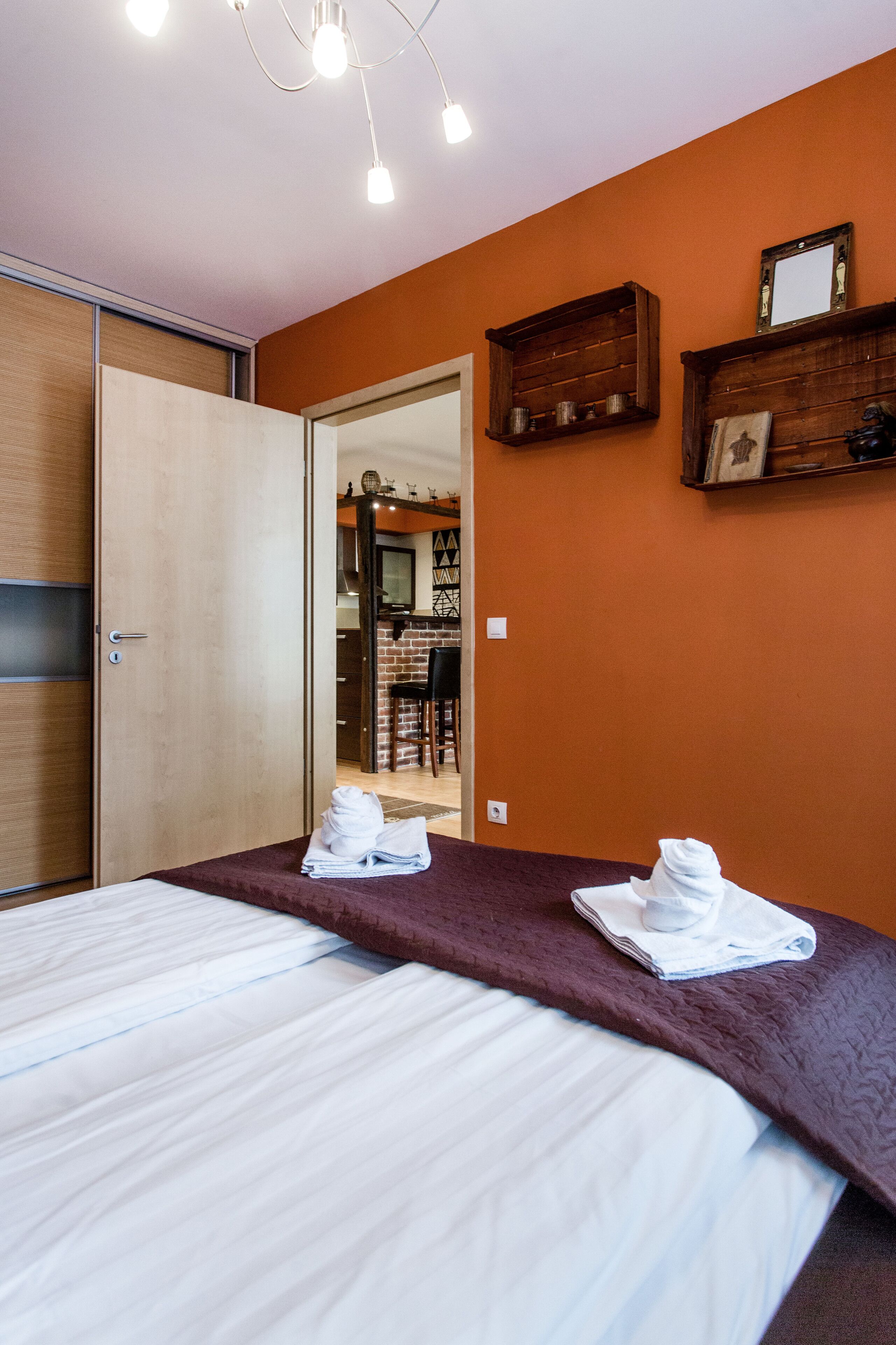 A hotel room with a bed, a wooden wardrobe, a mirror, and a small shelf with decorative items.