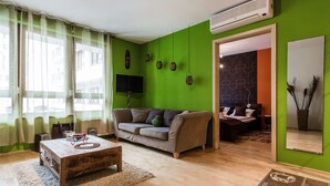 Ein Wohnzimmer mit grüner Wand, grauem Sofa, Couchtisch aus Holz und Flachbildfernseher.