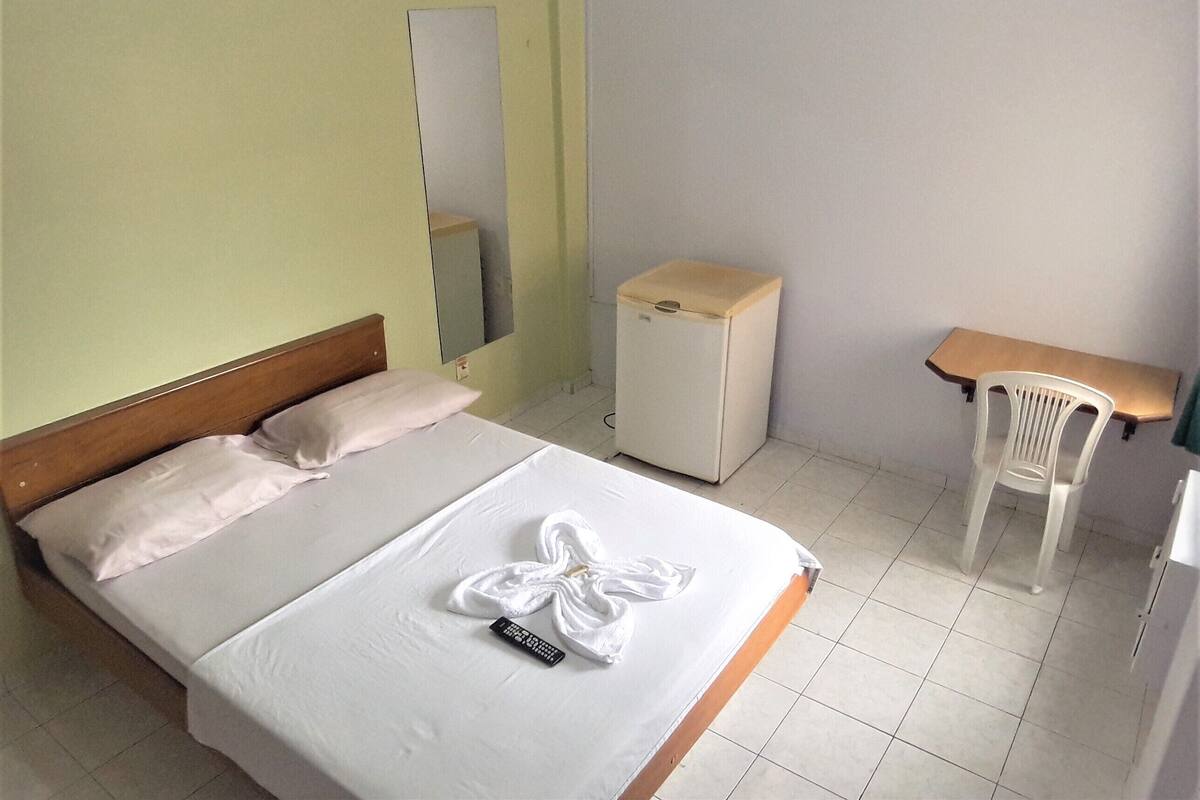 A hotel room with a bed, a small desk, a chair, a refrigerator, and a mirror.
