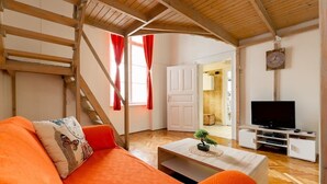 A cozy living room with a wooden ceiling, a sofa with orange cushions, a coffee table with a small plant, a television on a white stand, and a staircase leading to another room. - Hunyadi 3 Apartment (Budapest)