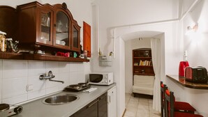 Cocina compacta con microondas integrado, lavamanos y un refrigerador pequeño. Hay un armario de madera sobre la encimera y una estantería en la habitación contigua.