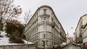 Un edificio histórico con arquitectura clásica, una pared de piedra y coches aparcados a lo largo de una calle nevada.