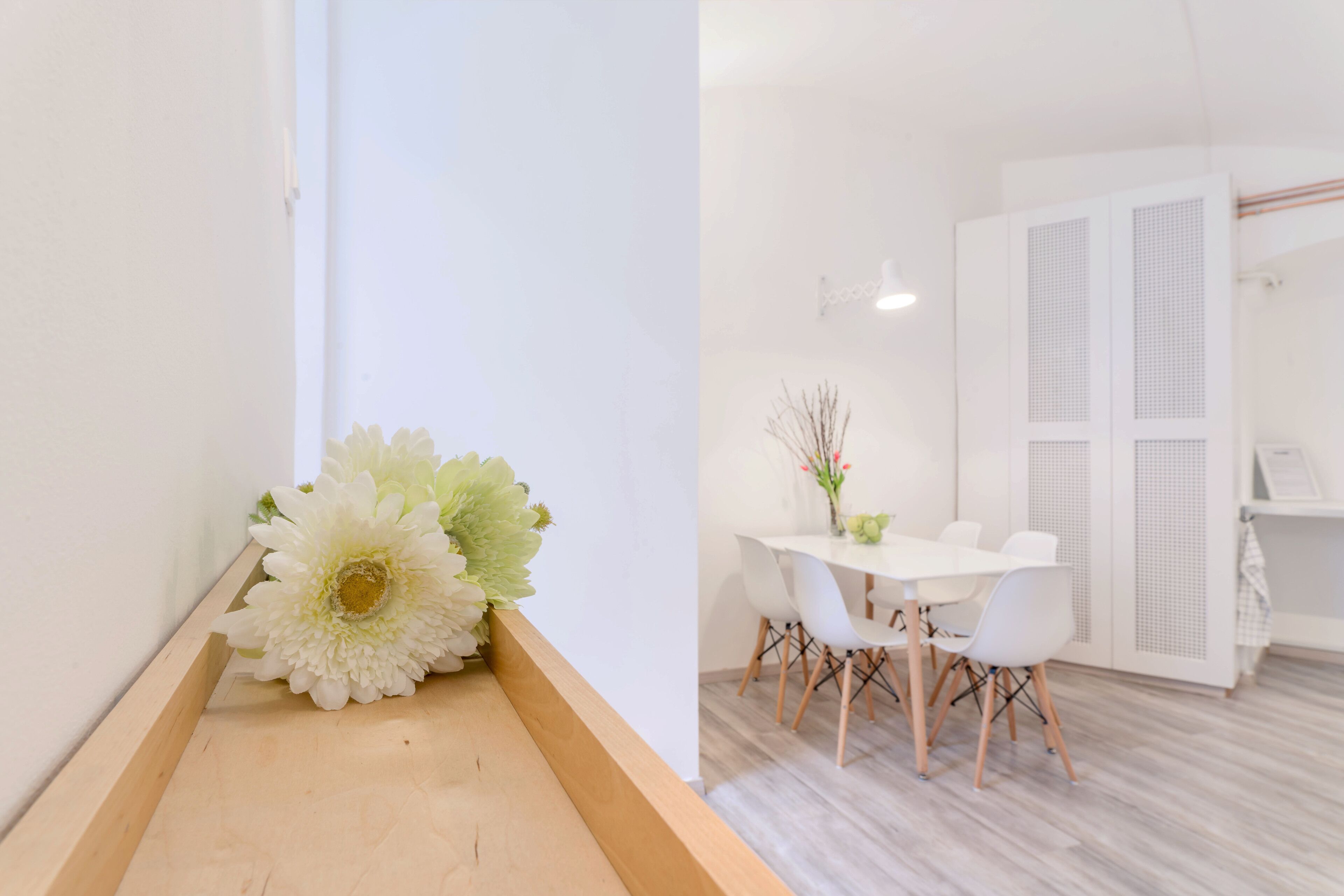 Ein modernes Interieur mit einer hölzernen Bank, weißen Wänden, einem Essbereich mit weißem Tisch und Stühlen sowie einem weißen Kleiderschrank.
