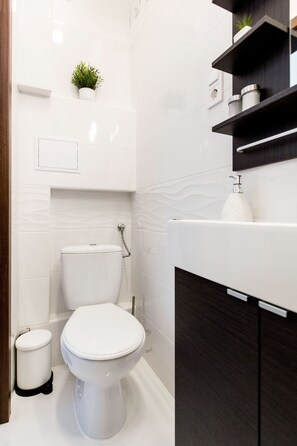 Een moderne badkamer met een wit toilet, een klein afvalbakje, een plank met toiletartikelen en een plant in een pot aan de muur.