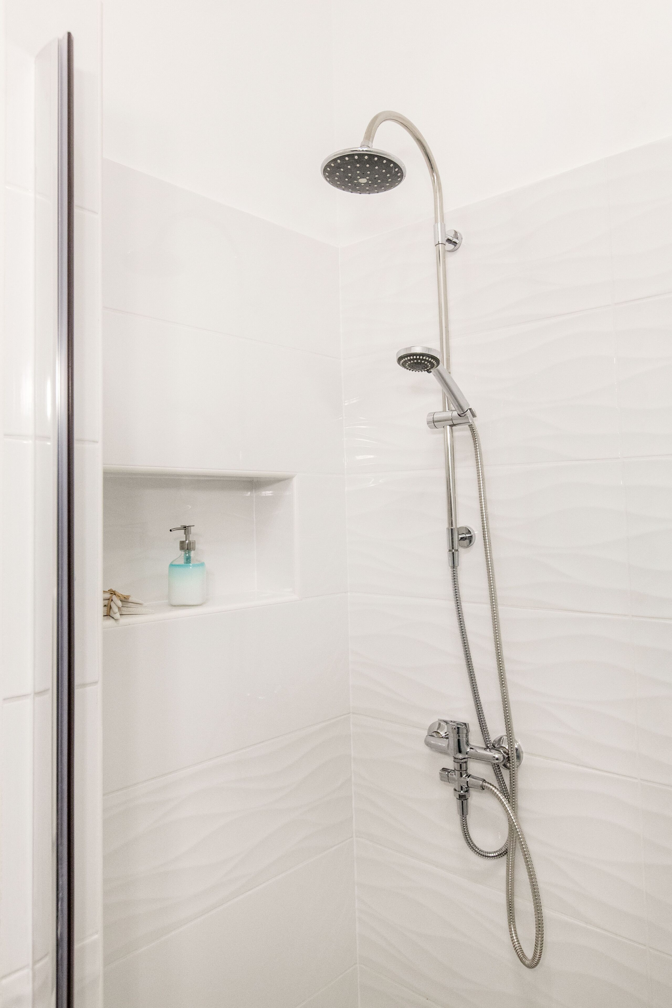 A shower with a handheld showerhead and a handheld hose, a soap dispenser on a shelf, and a shower control unit.