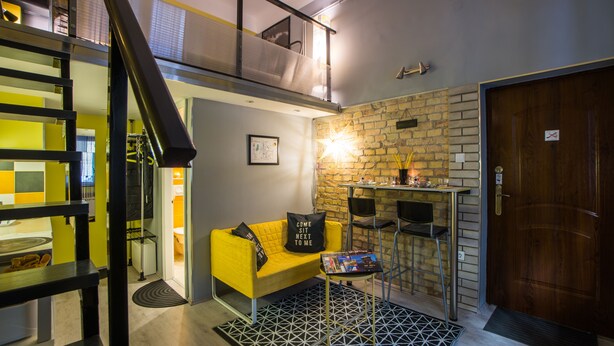 Ein moderner Wohnraum mit einem gelben Sofa, einem kleinen Tisch und einer Treppe, die in eine obere Ebene führt.