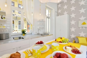 Une cuisine lumineuse avec des meubles blancs, une table à manger agrémentée de sets de table jaunes et un coin petit-déjeuner avec des fruits et des viennoiseries.
