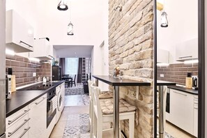 A modern kitchen with white cabinetry, a black countertop, and a stone accent wall. There is a dining area with a table and chairs, and a view into another room.