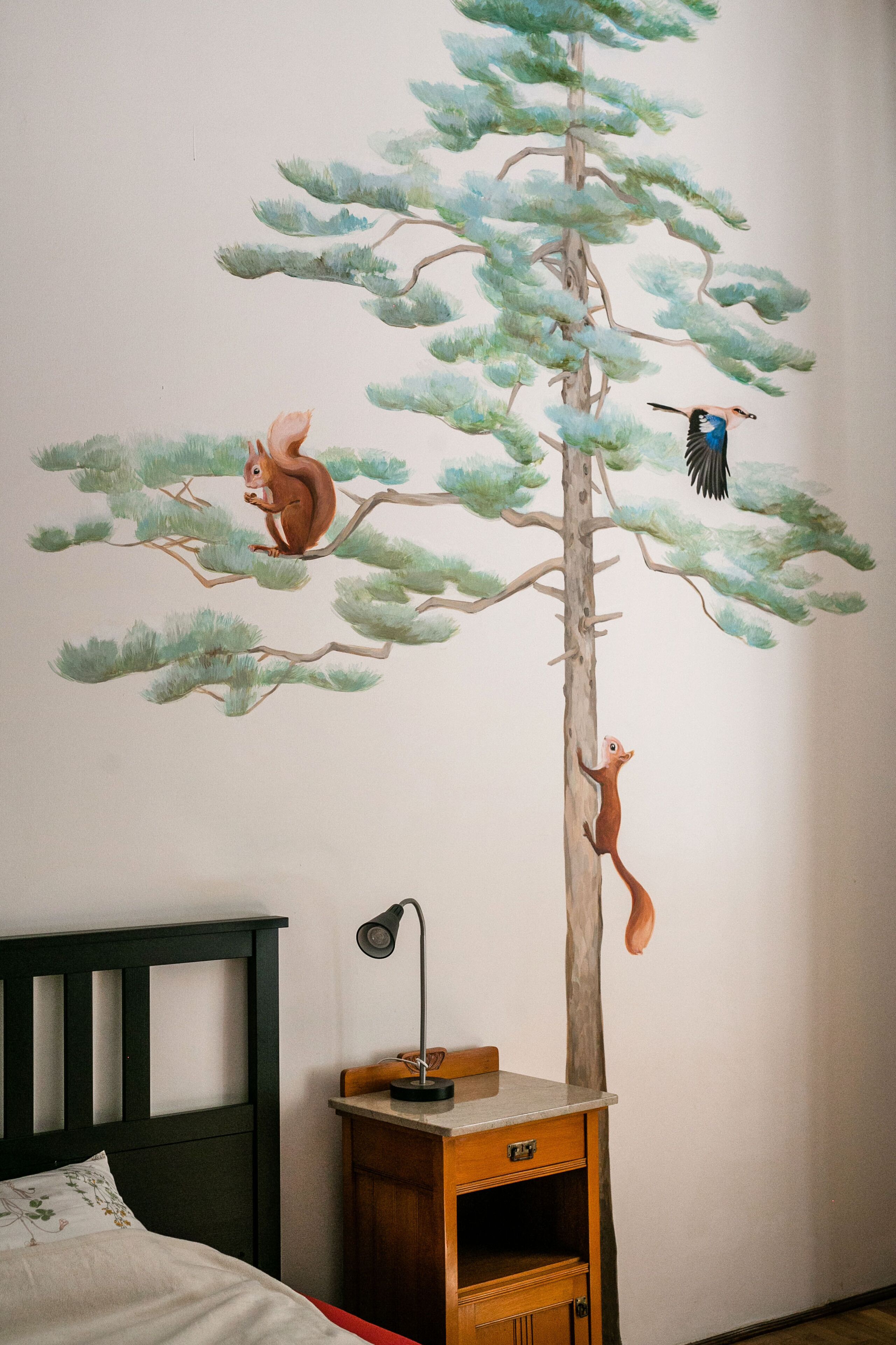 Un dormitorio con cama, mesita de noche y un mural de un árbol con ardillas.