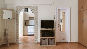 Una habitación con una televisión sobre un soporte de madera, una cocina visible a través de un arco y un armario.