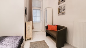 A small, well-lit room with a bed, a sofa, a radiator, and a door leading to another room. - Nagy Diofa 18 Apartment II. (Budapest)