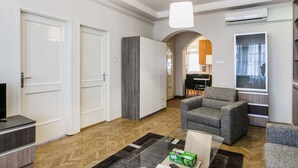 A modern living room with a grey sofa, a glass coffee table, and a TV. There is a wooden herringbone floor, white doors, and a kitchen visible in the background. - Nagymezo 33 Apartment (Budapest)