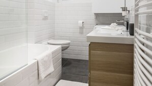 A modern bathroom with white tiles, a bathtub, a toilet, and a wooden vanity. - Nyar 7 Apartment (Budapest)