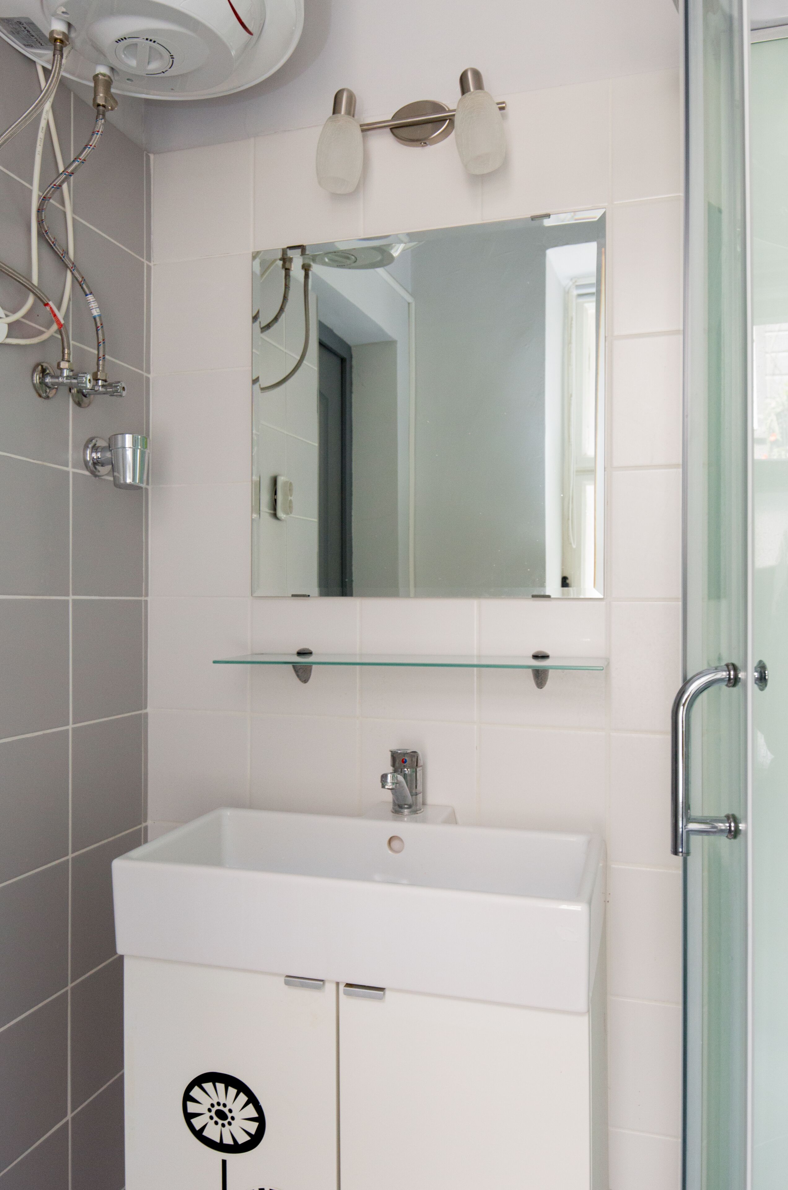 A bathroom with a white sink, a glass shelf above it, a mirror, and a showerhead.