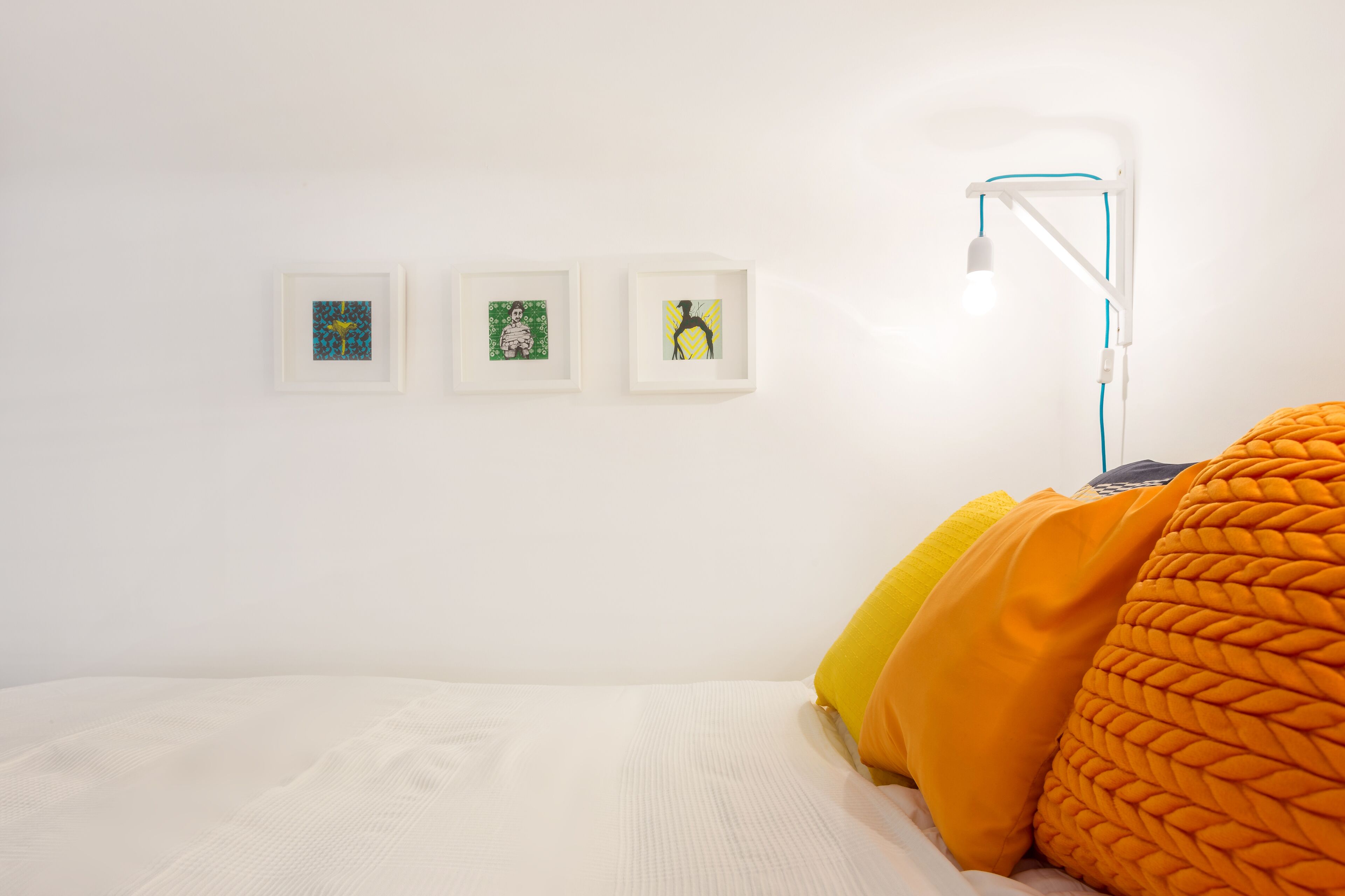 A bedroom with a white bed, three framed artworks on the wall, and a bedside lamp.