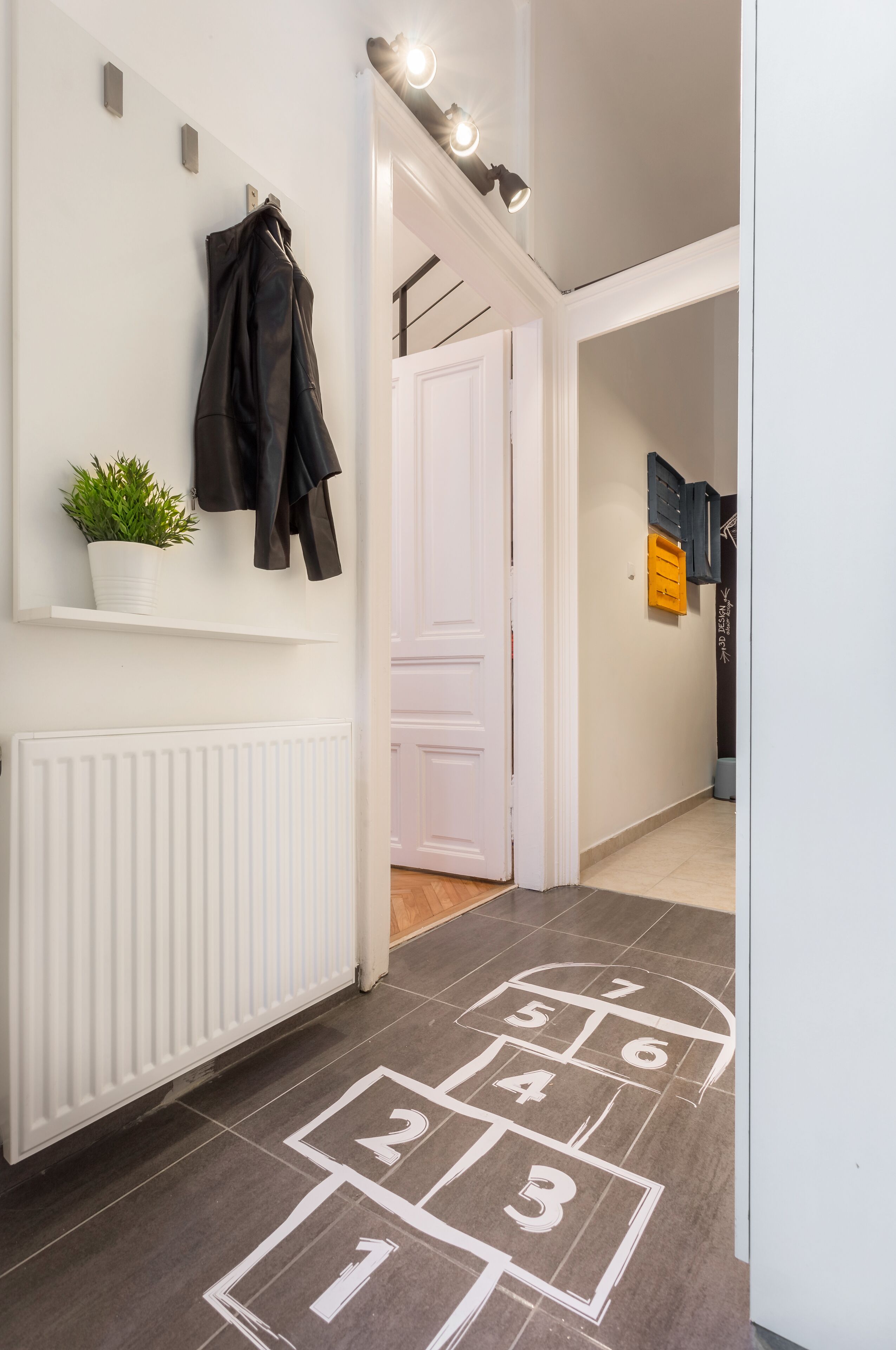 Un couloir avec un radiateur blanc, une plante en pot et un porte-manteau. On y trouve une porte sur laquelle est apposée une pancarte écrite dans une langue étrangère.
