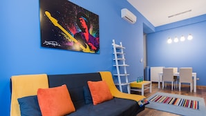 A modern living room with a blue wall, a yellow and black sofa, a colorful painting, a white ladder, a dining area with chairs and a table, and a blue rug. - Paulay 7 Apartment I. (Budapest)
