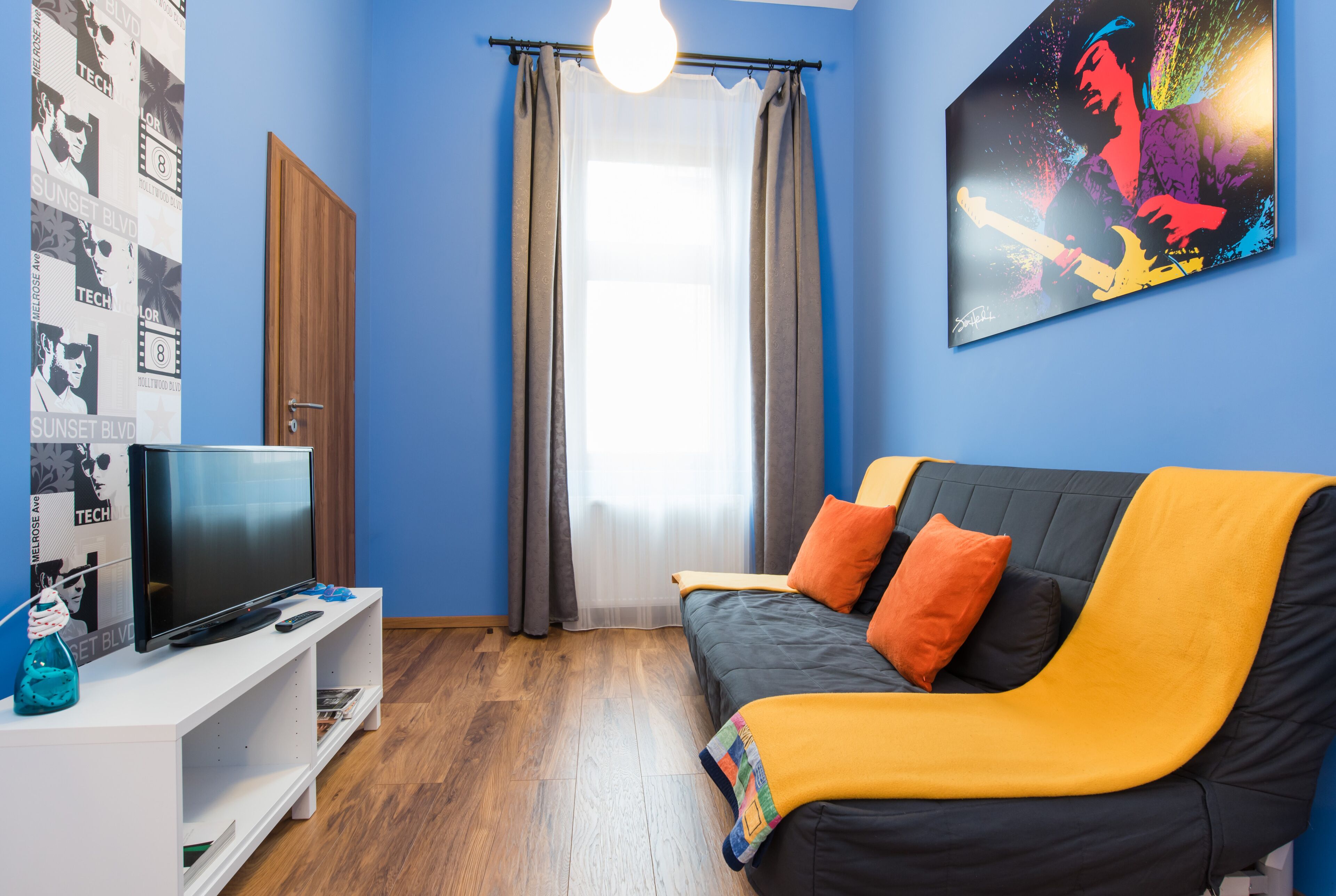 Ein Zimmer mit Fernseher auf einem weißen Schrank, einer Couch mit orangefarbenen und schwarzen Kissen, einer blauen Wand und einem Poster eines Musikers.