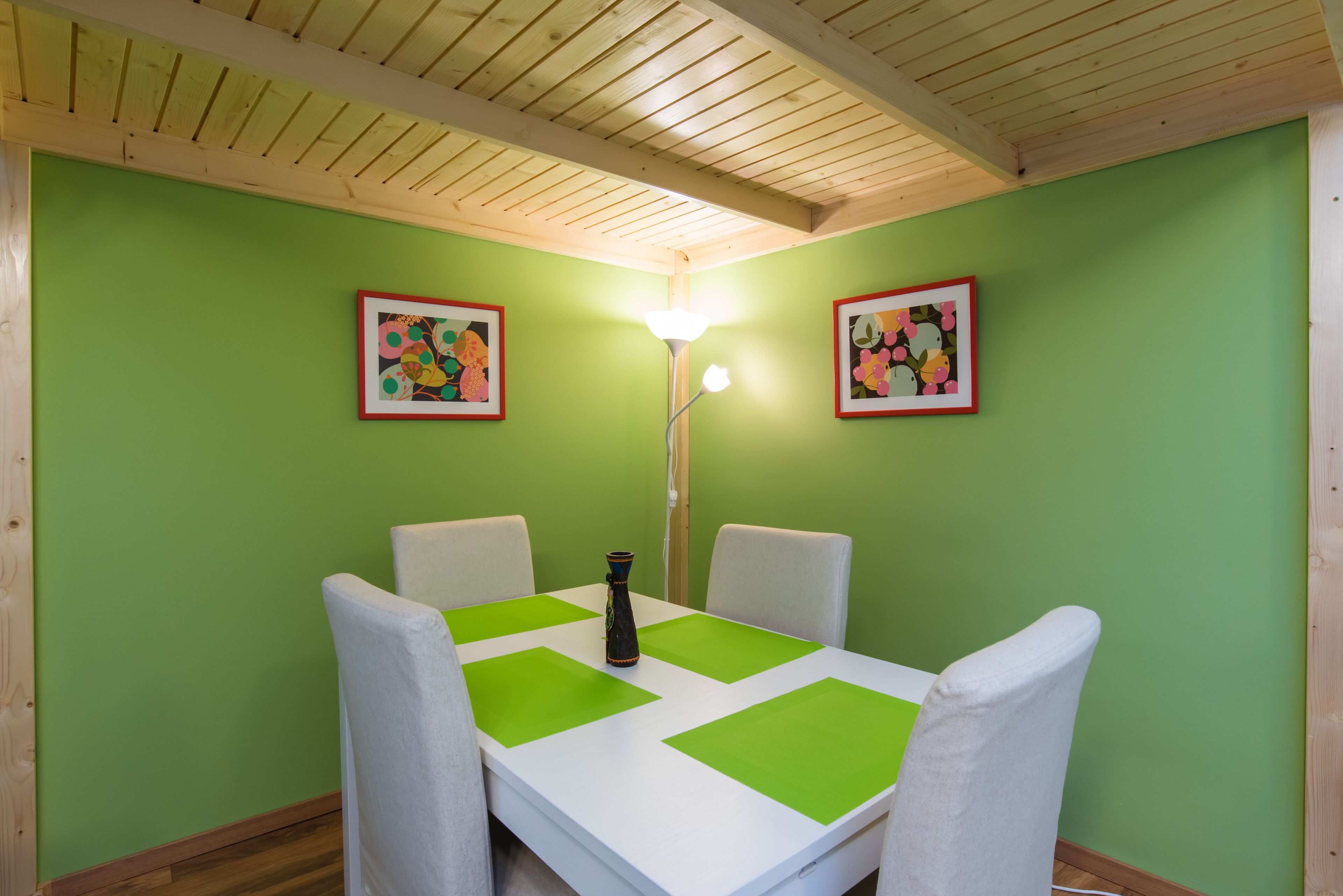 A dining area with a white table, green placemats, white chairs, and two framed artworks on green walls.