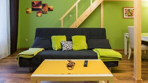 Sala de estar con pared verde, sofá negro con cojines verdes y negros, mesa de centro amarilla con un control remoto y un peluche, y una escalera de madera.