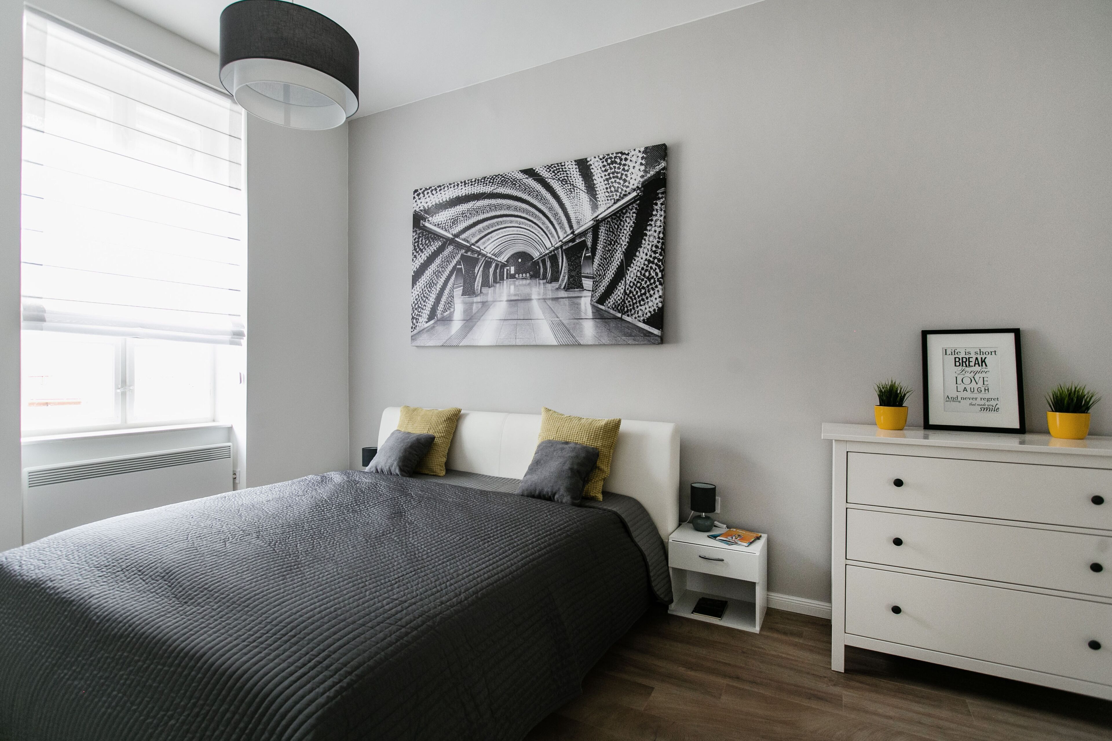 A modern bedroom with a large bed, a white dresser, a nightstand, and a wall-mounted abstract artwork.