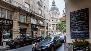 Una calle con coches aparcados, una mujer caminando y un edificio con una cúpula y un cartel que enumera alimentos.