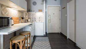 Cocina compacta con microondas, tostadora y taburetes de madera. Se ven un reloj y una lámpara en la pared.
