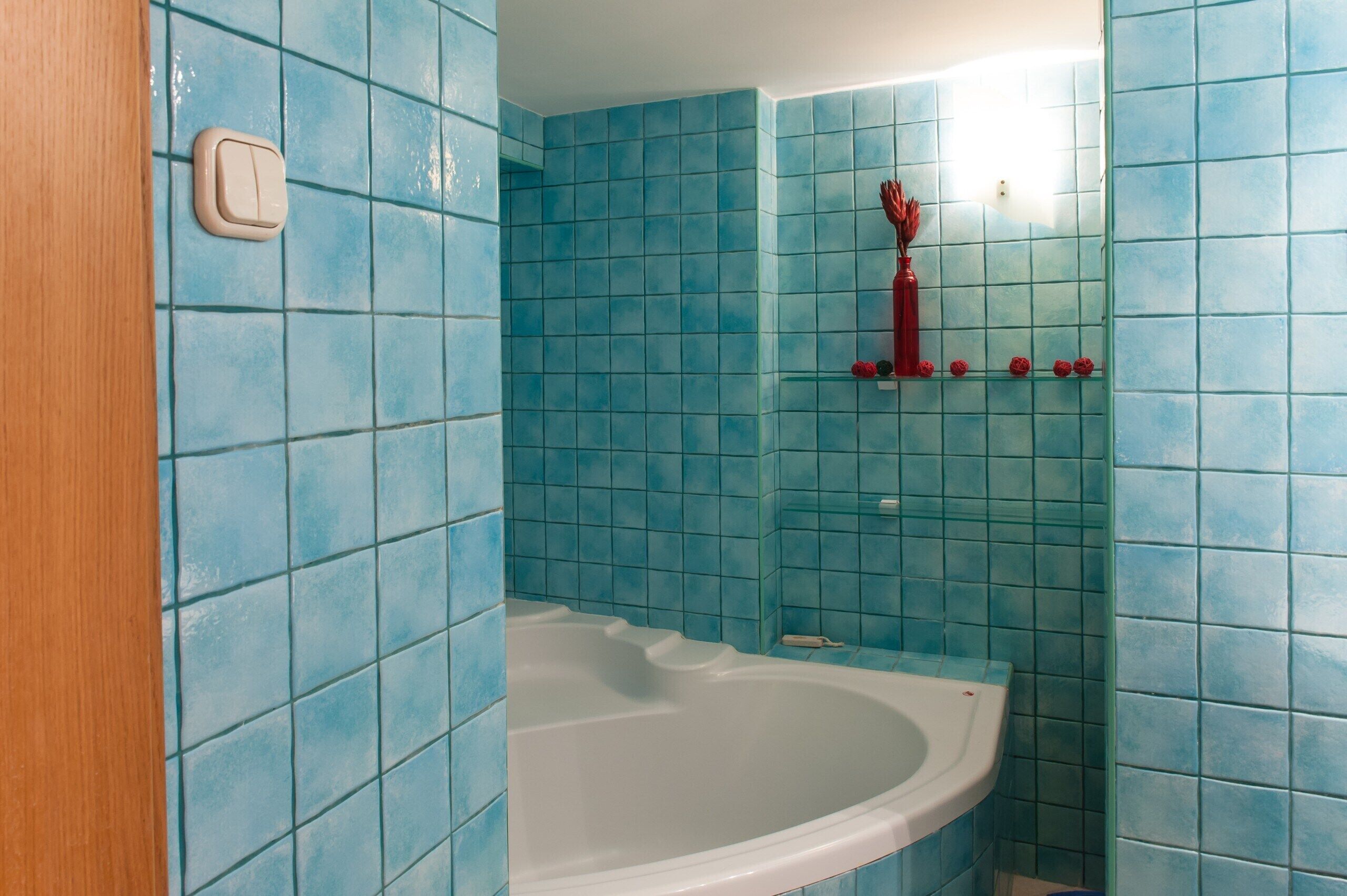 A bathroom with blue tiles, a white bathtub, and a red vase on a shelf.